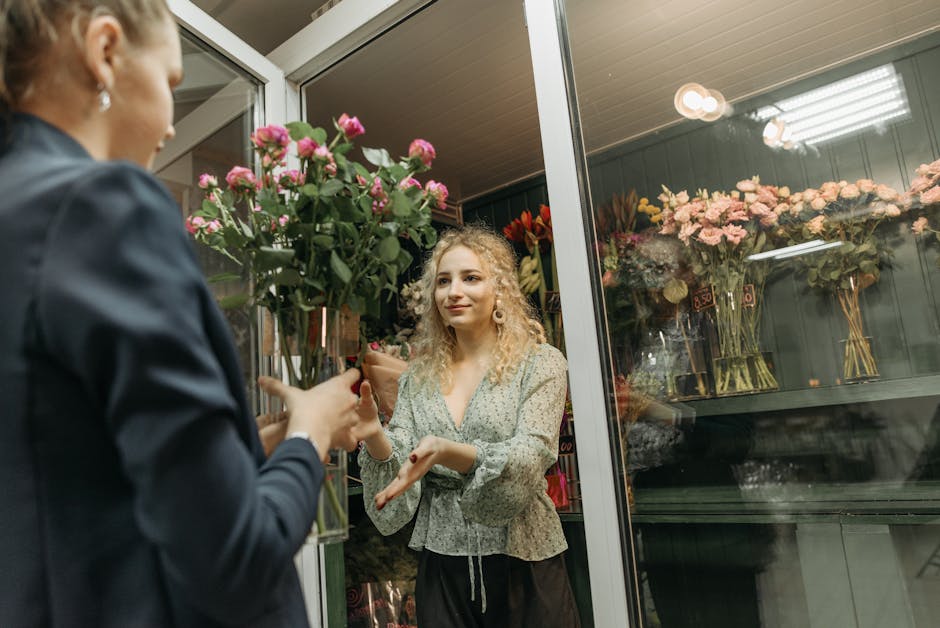 A florist from flowerschessington.co.uk, dressed in a dark jacket, hands a large bouquet of fresh pink roses to a young woman with curly blonde hair, wearing a patterned blouse, inside a flower shop.