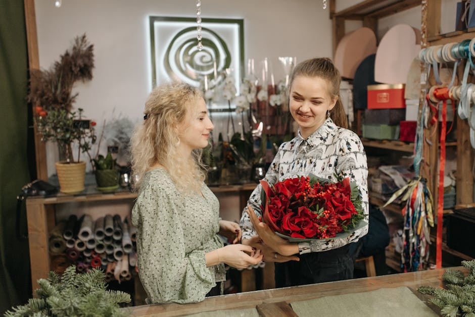 A young woman with light brown hair tied back, wearing a patterned shirt, is smiling while holding a bouquet of fresh, vibrant red roses, mixed with red carnations and greenery, wrapped in dark green