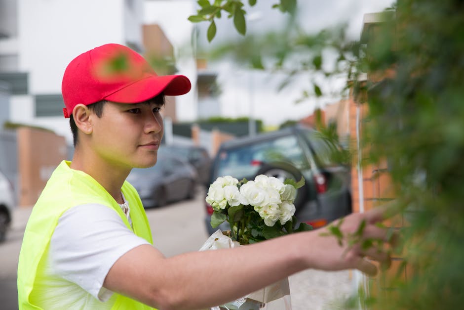A young male florist wearing a red cap and a bright yellow safety vest arranges a bouquet of fresh white flowers, likely hydrangeas or roses, against a background of a residential street with parked c