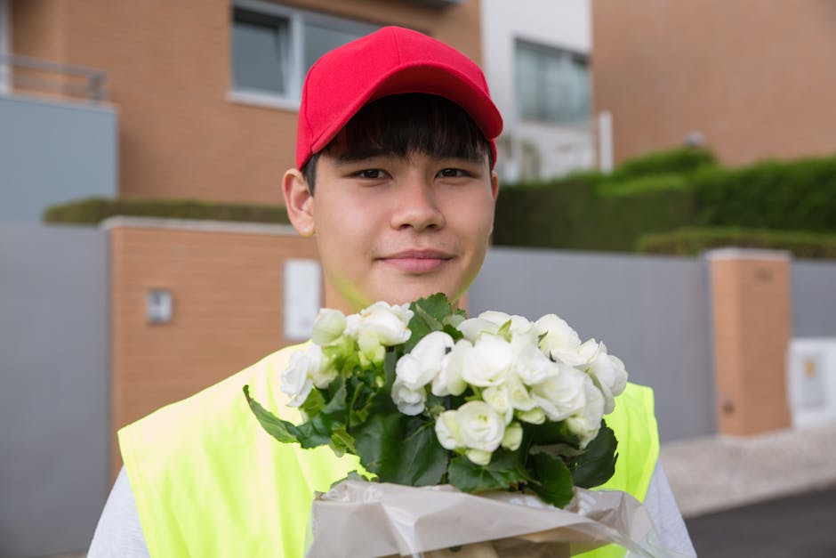 A young delivery person with light skin, dark hair, and a gentle smile, wearing a bright yellow high-visibility vest and a red cap, is holding a bouquet of fresh white flowers wrapped in white paper.