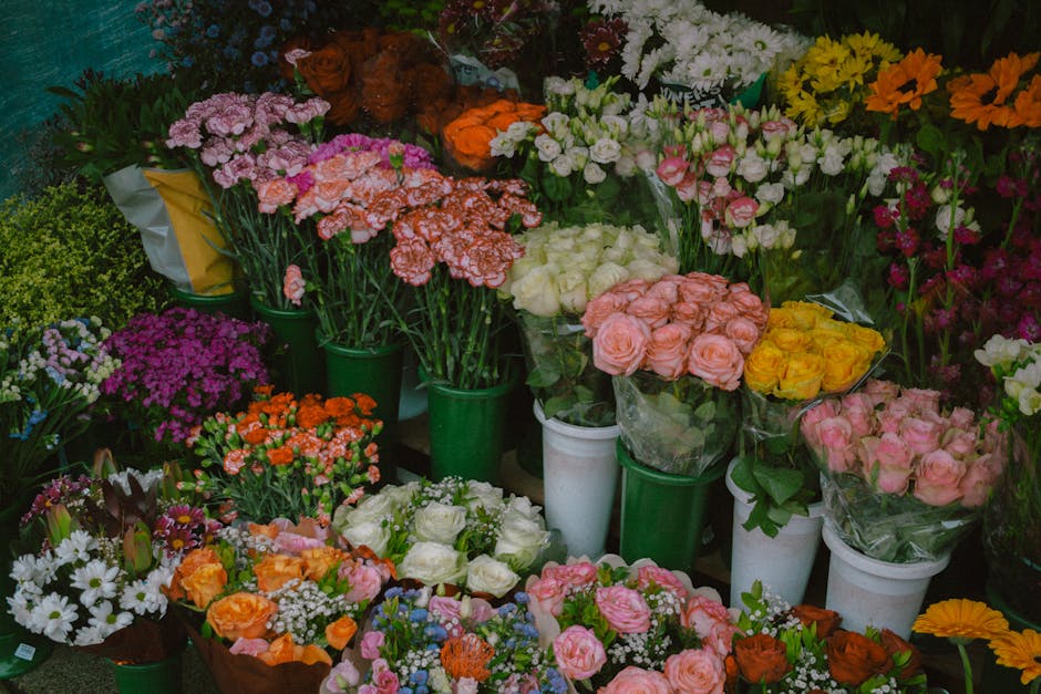 A vibrant display of fresh flowers arranged in various bouquets and containers, featuring pink, white, yellow, orange, purple, and red blooms. The flowers include roses, carnations, daisies, and other