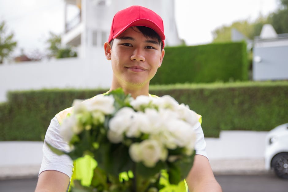 A delivery person wearing a bright yellow high-visibility vest and a red cap is standing outdoors, holding a bouquet of fresh white roses with green foliage. The bouquet is wrapped in clear plastic an
