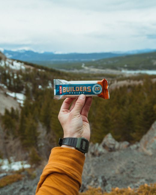 A close-up of a person's hand holding a protein bar labeled 'Builders 20', with a rugged outdoor landscape in the background featuring rolling hills, sparse trees, and distant mountains under a partly