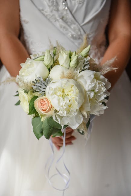 A bride dressed in a white wedding gown holds a round bouquet arrangement featuring an assortment of fresh flowers, including soft white peonies, delicate pale pink roses, and white lisianthus, comple